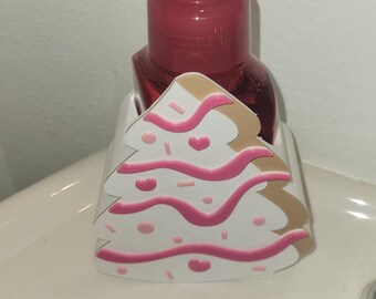 Könnte beinhalten: Ein rosa Seifenspender mit einem dekorativen, weiß-rosa Weihnachtsbaum-förmigen Halter. Der Spender steht auf einem weißen Waschbecken. Der Spender hat eine rosa Pumpe und eine transparente Flasche.