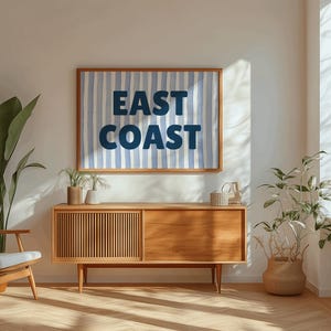 May include: A wooden mid-century modern style cabinet with two doors and a drawer. The cabinet is in a living room with a blue and white striped poster that says "EAST COAST" hanging on the wall above it. There are two potted plants on either side of the cabinet.