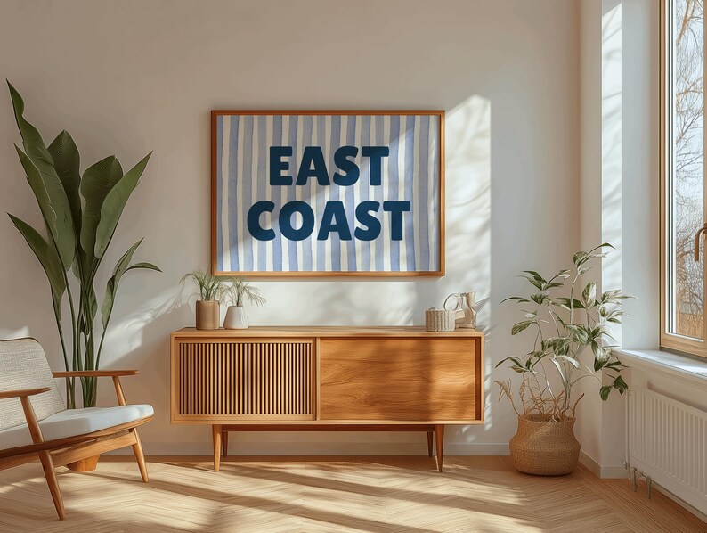 May include: A wooden mid-century modern style cabinet with two doors and a drawer. The cabinet is in a living room with a blue and white striped poster that says "EAST COAST" hanging on the wall above it. There are two potted plants on either side of the cabinet.