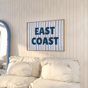 May include: A white couch with two pillows and a framed print on the wall behind it. The print has a blue and white striped background with the text "EAST COAST" in dark blue.