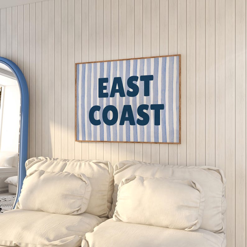 May include: A white couch with two pillows and a framed print on the wall behind it. The print has a blue and white striped background with the text "EAST COAST" in dark blue.