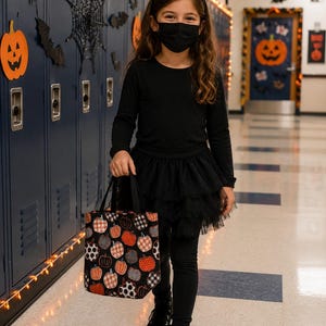 A black tote bag featuring a repeating pattern of orange, white, and black pumpkins in various plaid and polka dot designs. The bag has two black handles and is constructed from fabric, showcasing a festive seasonal aesthetic.