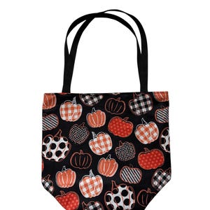 Op de afbeelding: Een zwarte draagtas met een herhalend patroon van pompoenen in verschillende tinten oranje, wit en zwart. De pompoenen zijn versierd met diverse prints, waaronder ruitjes, stippen en strepen. De tas heeft twee zwarte stoffen handvatten.