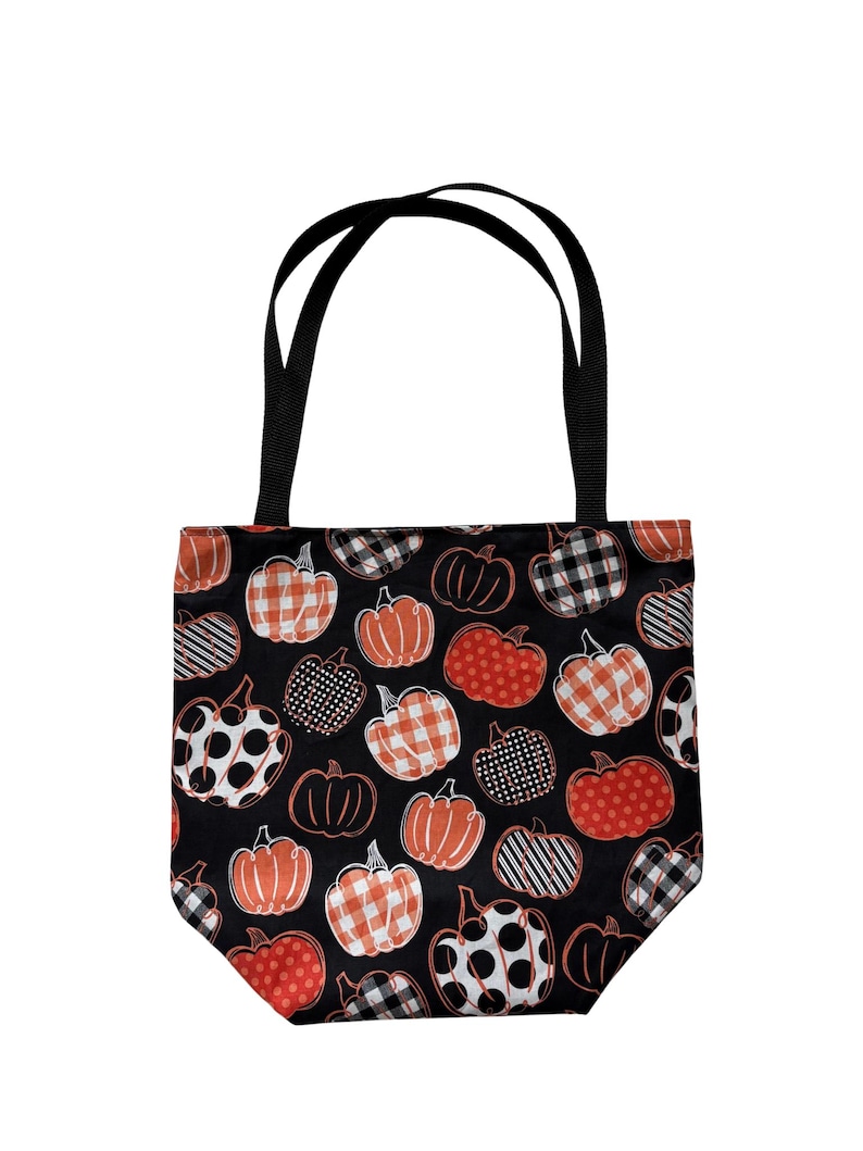 Op de afbeelding: Een zwarte draagtas met een herhalend patroon van pompoenen in verschillende tinten oranje, wit en zwart. De pompoenen zijn versierd met diverse prints, waaronder ruitjes, stippen en strepen. De tas heeft twee zwarte stoffen handvatten.