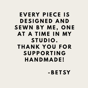 May include: A rectangular sign with a light beige background and black text that reads: EVERY PIECE IS DESIGNED AND SEWN BY ME, ONE AT A TIME IN MY STUDIO. THANK YOU FOR SUPPORTING HANDMADE! -BETSY. The text is centred and in a sans-serif font.