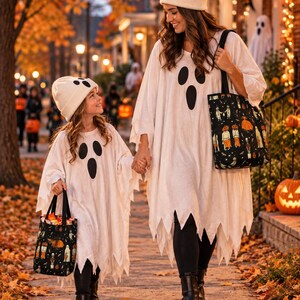 Two white ghost costumes featuring black oval eyes and mouth prints, paired with matching cream-colored beanies. Each costume includes a black tote bag decorated with a repeating pattern of pumpkins and figures, worn over black leggings.