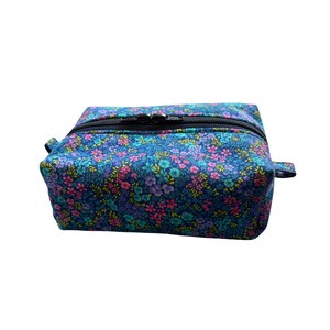 A rectangular fabric cosmetics bag with a floral print in shades of blue, pink, and yellow, featuring a black zippered closure across the top.