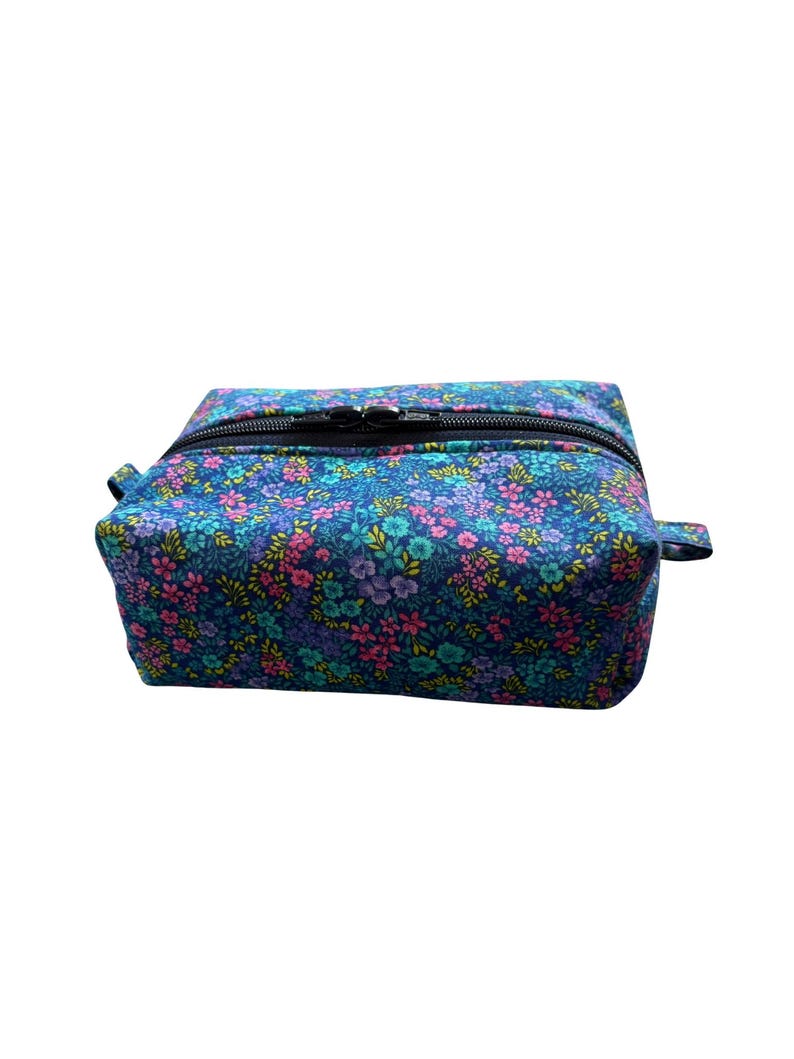 A rectangular fabric cosmetics bag with a floral print in shades of blue, pink, and yellow, featuring a black zippered closure across the top.