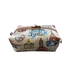 A rectangular fabric cosmetics pouch featuring a colorful sketch print of famous landmarks and a brown zippered closure across the top.