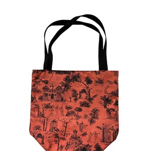 Op de afbeelding: Een draagtas met een roestoranje achtergrond en een zwarte toile-print met diverse architecturale structuren, bomen en kleine figuren. De tas heeft twee zwarte stoffen hengsels aan de bovenkant om de tas aan te dragen.