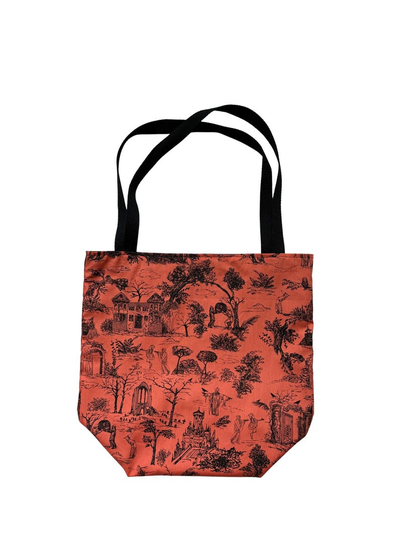 Op de afbeelding: Een draagtas met een roestoranje achtergrond en een zwarte toile-print met diverse architecturale structuren, bomen en kleine figuren. De tas heeft twee zwarte stoffen hengsels aan de bovenkant om de tas aan te dragen.
