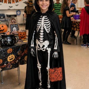 A black hooded cape worn over a black jumpsuit featuring a white skeleton print on the front. The costume includes matching skeleton-printed gloves and is paired with black and white sneakers while holding a small orange patterned bag.