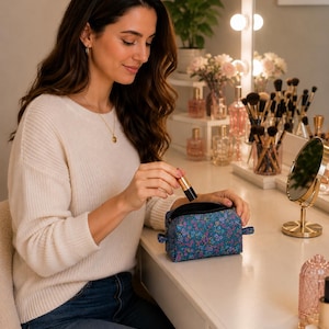 A smiling woman sits at a vanity table, holding a small lipstick next to an open blue floral-patterned zippered cosmetics pouch.