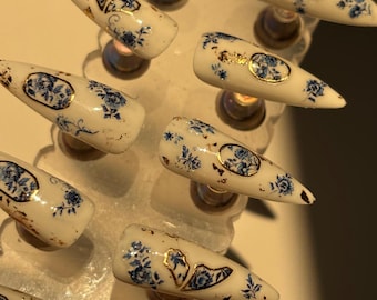May include: Set of ten white false nails with a blue and gold floral design. The nails are arranged on a white display stand.
