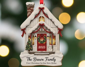 Puede incluir: Adorno navideño en forma de casa blanca con puerta roja, tejado nevado y pequeño árbol de Navidad. El adorno tiene el texto "2025" y "The Brown Family" con "First Christmas In Our New Home". Se sujeta con una cinta roja.