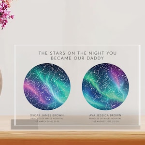May include: A clear acrylic block displays a personalized star map with the text "THE STARS ON THE NIGHT YOU BECAME OUR DADDY." Two circular star maps are featured, with names and dates below. The background includes a vase of flowers and a clock.