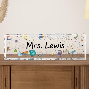 May include: A rectangular clear acrylic block desk sign featuring the text Mrs. Lewis in black font. The surface is decorated with colorful science-themed illustrations including a prism, lightbulb, telescope, calculator, and physics book.