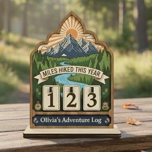 May include: Wooden adventure log with a mountain and river scene, displaying "MILES HIKED THIS YEAR" and the number "123". The log has a decorative top with a sun and mountain design. The text "Olivia's Adventure Log" is at the bottom.