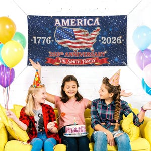 Puede incluir: Una pancarta con la palabra "AMERICA" y los años "1776" y "2026" está en exhibición. La pancarta también presenta la bandera estadounidense en forma de los Estados Unidos. Tres chicas están sentadas en un sofá amarillo con globos y un pastel.