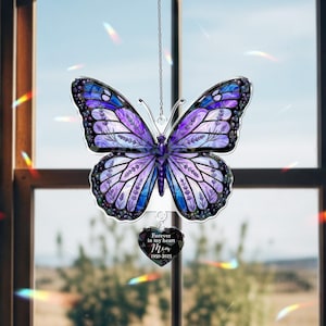 May include: A hanging butterfly ornament featuring purple and blue stained glass patterns with black outlines. A small heart-shaped pendant hangs from the bottom, inscribed with the text Forever in my heart Mom 1950-2025 in white lettering.