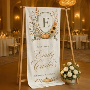 Op de afbeelding: Een verticaal stoffen spandoek op een houten ezel met een centraal schild met de letter E, omringd door herfstbladeren en pompoenen. De tekst luidt: Welcome to Emily Carter's Bridal Shower, Saturday, October 18, 2025, in gouden letters.