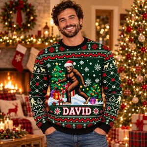 May include: A dark green long-sleeve jumper featuring a graphic of a brown Bigfoot wearing a Santa hat, standing next to Christmas trees and wrapped gifts. The jumper includes white snowflake patterns and the name DAVID in white block letters.