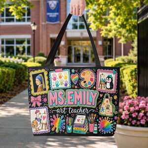 Puede incluir: Un bolso tote negro con un diseño artístico colorido y el texto "MS. EMILY art teacher". El estampado incluye ilustraciones de una pintura, un pingüino, un ganso, huellas de manos, un corazón arcoíris, pinceles y un cuaderno de bocetos.