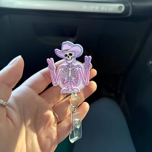 Puede incluir: Un portatarjetas con un esqueleto que lleva un sombrero de vaquero de color morado claro. El esqueleto se muestra con ambas manos levantadas haciendo el signo de la paz. El diseño utiliza tonos rosa, morado y blanco, con un clip de plástico.