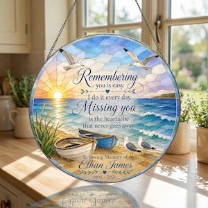 May include: A circular stained glass suncatcher featuring a coastal scene with two boats on a sandy beach, two seagulls flying above, and two seagulls standing on the sand. The text reads: Remembering you is easy. I do it every day. Missing you is the heartache that never goes away. In Loving Memory of Ethan James 1995 - 2026.