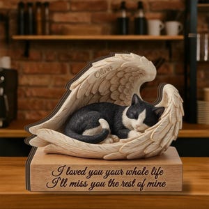 Puede incluir: Una figura conmemorativa de un gato blanco y negro durmiendo, acurrucado en alas de ángel esculpidas. Las alas son de color crema y la base es de color madera. El nombre "Oliver" está inscrito en las alas. La base dice: "Te amé toda tu vida, te extrañaré el resto de la mía."