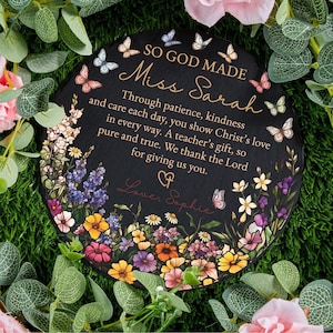 Puede incluir: Placa redonda de pizarra negra con diseños florales y mariposas. La placa tiene el texto "SO GOD MADE Miss Sarah" y un mensaje de agradecimiento. La placa está rodeada de vegetación y flores.