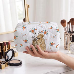 May include: A white quilted cosmetic bag featuring a floral pattern with orange and blue flowers. The bag displays a printed illustration of a brown cat and a golden retriever dog, with the names Beth and Cookie written in black script text.