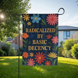 Op de afbeelding: Een marineblauwe tuinvaandel met kleurrijke bloemmotieven en de gouden tekst "RADICALIZED BY BASIC DECENCY". De vlag wordt getoond op een zwarte metalen standaard in een tuin.