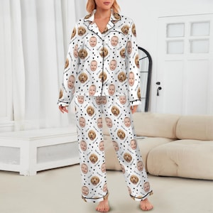 May include: White pajama set with a black trim, featuring a pattern of baby faces, brown dogs, and black hearts. The long-sleeved top has a collared neckline and button-down front. The matching pants have an elastic waistband.