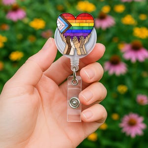 May include: A retractable badge reel featuring a heart-shaped design with the LGBTQ+ pride flag and hands. The heart is composed of rainbow-colored bricks. The reel has a clear plastic body and a metal clip.