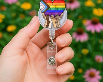 May include: A circular badge reel featuring a heart-shaped graphic with a brick-patterned pride flag design. The heart is held up by multiple illustrated hands of varying skin tones. The badge reel has a clear plastic strap with a metal snap button.