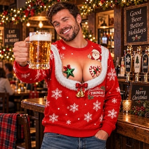 Può includere: Una felpa rossa a maniche lunghe con scollo in pelliccia sintetica bianca, una grafica di Babbo Natale, una stella verde, un bastoncino di zucchero e due campanelle dorate. La felpa riporta il nome THOMAS stampato in un riquadro bianco.