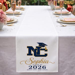 May include: A white fabric table runner featuring a navy blue and gold glitter graphic with the letters NC, a graduation cap icon, the name Sophia in gold script, and the text Class of 2026 North Coast High School printed on the end.