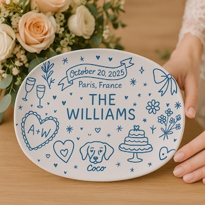 May include: White ceramic oval plate with blue illustrations and text. The plate features a banner with "October 20, 2025, Paris, France" and "THE WILLIAMS". Decorative elements include hearts, stars, flowers, and a dog illustration.