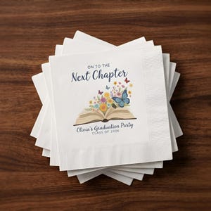 Op de afbeelding: Witte papieren servetten gestapeld op een houten oppervlak. Elke servet is bedrukt met de tekst "On to the Next Chapter" en "Olivia's Graduation Party CLASS OF 2026". Het ontwerp bevat een open boek met bloemen en vlinders.