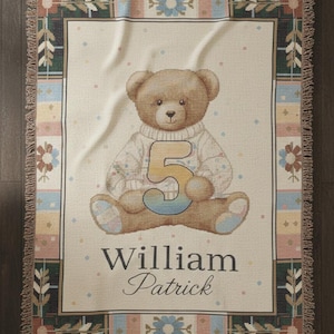 Pode incluir: Uma manta tecida com um ursinho de pelúcia vestindo um suéter com o número 5. A manta tem um fundo creme com uma borda decorativa em tons de verde, azul e rosa. O nome "William Patrick" está escrito abaixo.