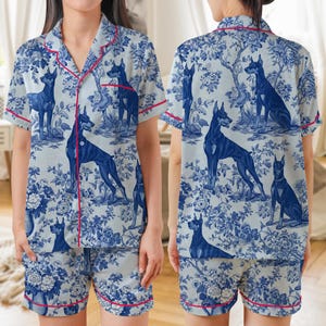 May include: A short-sleeved button-down pajama shirt and matching shorts set in a light gray fabric featuring a blue toile-style print of Doberman dogs and floral patterns, accented with thin red piping along the collar, placket, and hems.