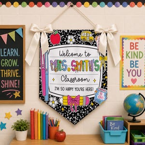 Könnte beinhalten: Ein hängendes Klassenzimmer-Banner mit schwarz-weiß gesprenkeltem Hintergrund und dem Text Welcome to MRS. SMITH'S Classroom I'M SO HAPPY YOU'RE HERE. Das Banner ist mit Grafiken eines roten Apfels, eines Schulbusses und eines Bleistifts verziert.