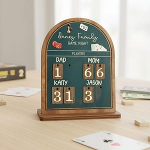 Könnte beinhalten: Eine Holz-Spielabend-Anzeigetafel mit tealfarbenem Hintergrund und den Worten "Innes Family Game Night". Die Anzeigetafel zeigt die Ergebnisse für "Dad" und "Mom" mit den Namen und Ergebnissen der einzelnen Spieler an. Dekorative Spielsteine befinden sich oben.