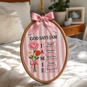 May include: An oval wooden embroidery hoop wall hanging featuring a pink and white striped fabric background. It displays the text GOD SAYS JAM and the name JAMIE vertically, with floral illustrations and five rectangular boxes containing scripture.