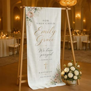 May include: A vertical white fabric banner with a subtle linen texture, featuring floral accents in the top left and bottom right corners. The text reads: FOR THIS CHILD Emily Grace We have prayed 1 SAMUEL 1:27 BAPTIZED 03.10.2026 with a gold cross.