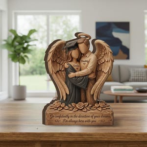 May include: A wooden angel sculpture featuring a father and daughter embracing, with angel wings and a halo. The daughter wears a graduation cap and gown. The sculpture is engraved with the names "Emily" and "Daddy", and the quote "Go confidently in the direction of your dreams, I'm always here with you."