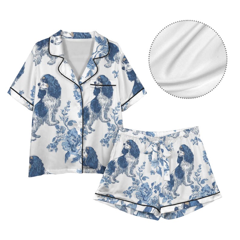 Puede incluir: Conjunto de pijama blanco con un estampado floral y de perros en azul y blanco. El conjunto incluye una camisa de manga corta con botones y pantalones cortos a juego, ambos con ribete negro. La tela parece sat&eacute;n.