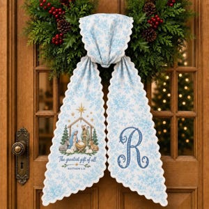 May include: A light blue and white floral fabric door hanger with scalloped edges. One side features a nativity scene with the text The greatest gift of all and MATTHEW 1:21. The other side displays a large, ornate blue letter R monogram.