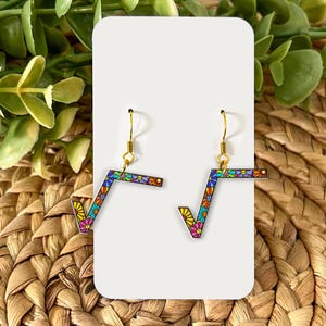 Puede incluir: Un par de pendientes con el símbolo de la raíz cuadrada y un colorido estampado floral. Los pendientes tienen ganchos dorados y se exhiben en una tarjeta blanca sobre un fondo de paja tejida y follaje verde.